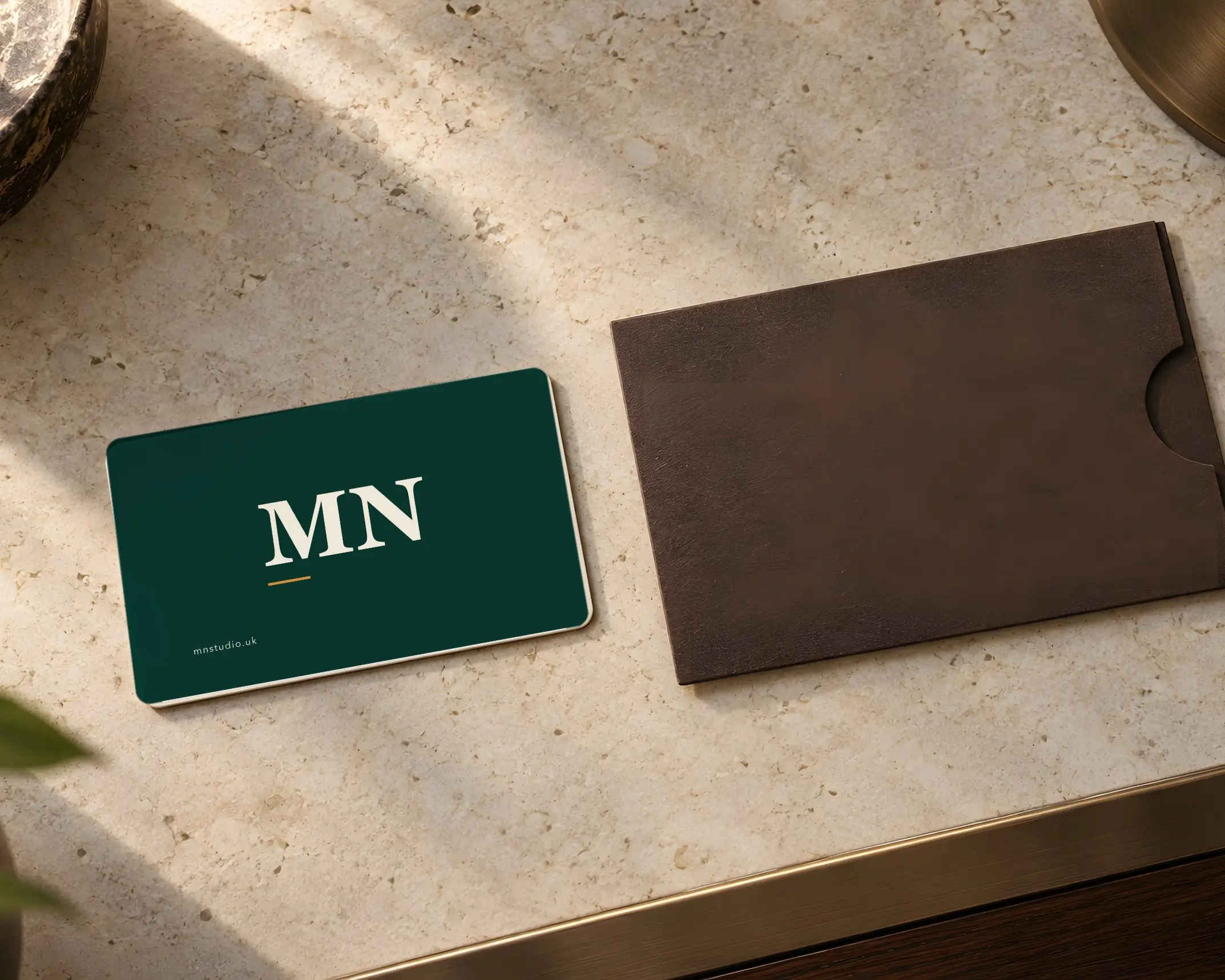 A green MN Studio business card with the MN monogram beside a brown leather card sleeve on a stone table