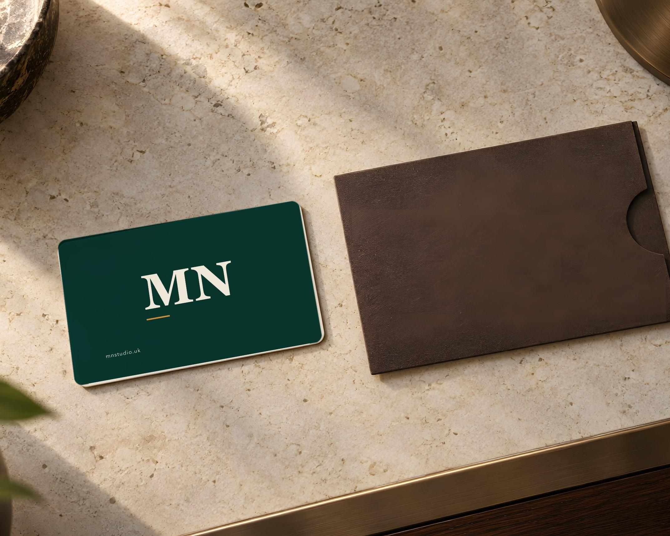 A green MN Studio business card with the MN monogram beside a brown leather card sleeve on a stone table