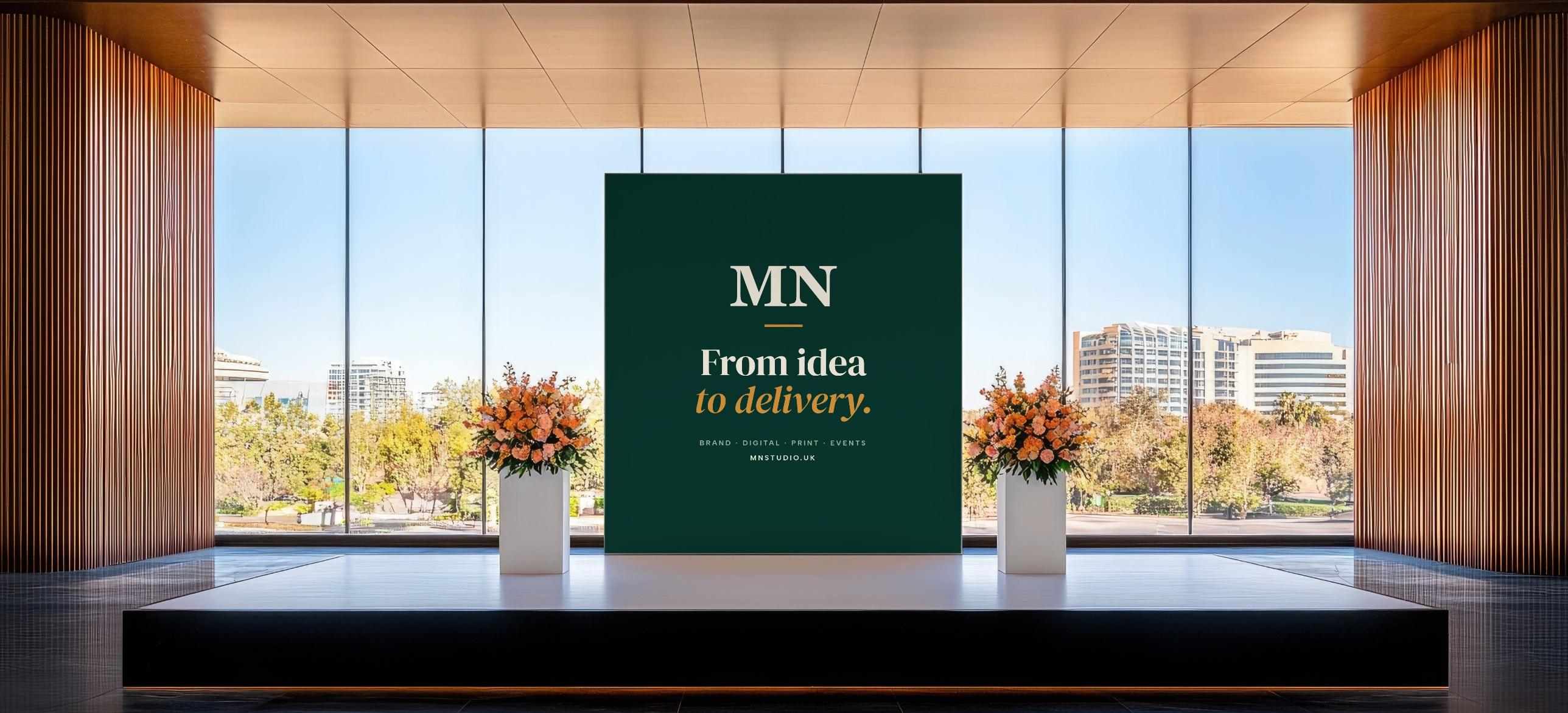 A stage in a bright glass-walled venue with a green MN Studio backdrop reading “From idea to delivery.” between two floral arrangements